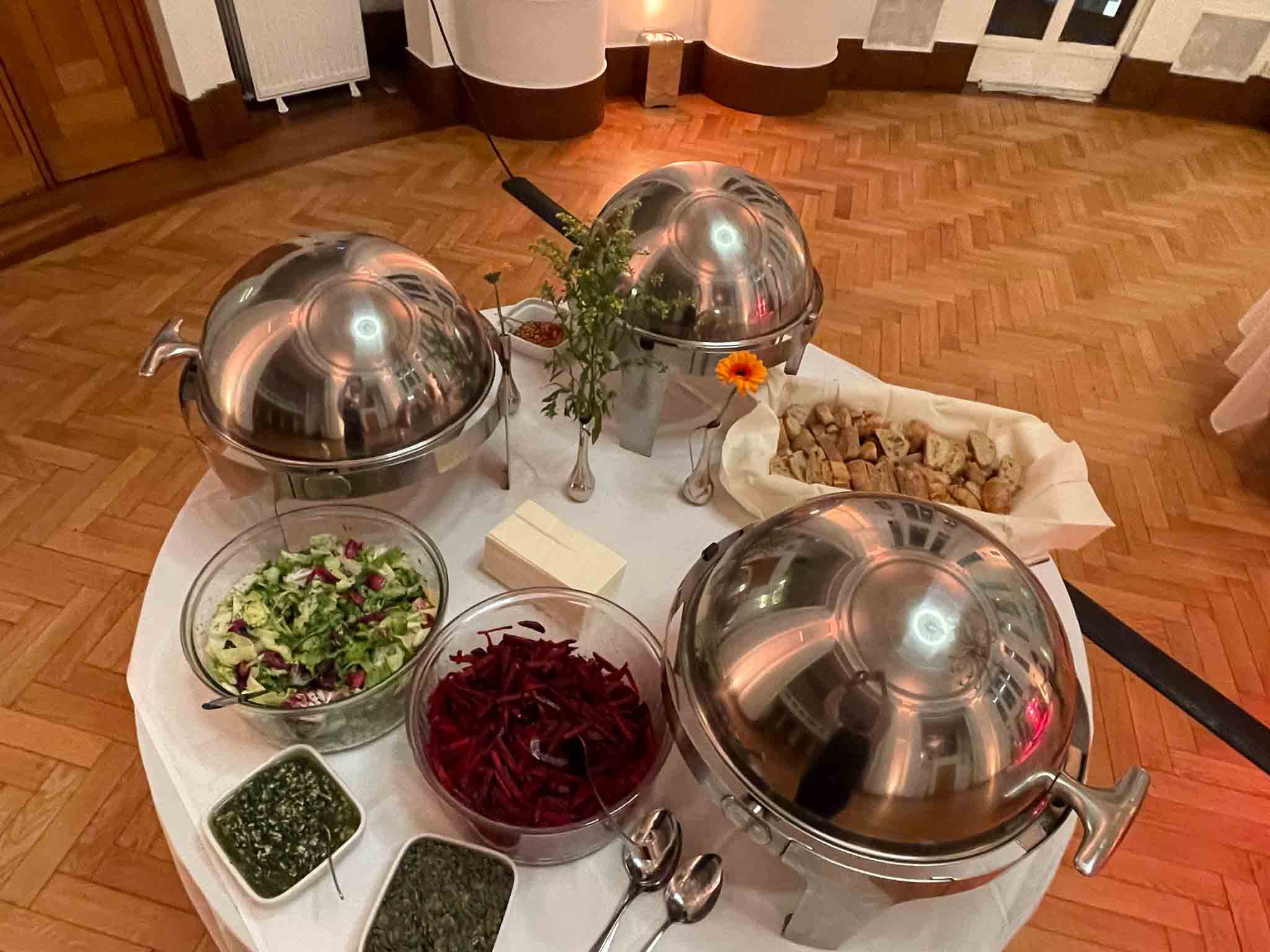 Een witte tafel met meerdere zilveren schalen met daarnaast een kom rodebietsalade, een kom komkommersalade en een broodmand.